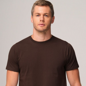 Portrait of a man with short light brown hair wearing a plain brown T-shirt against a neutral gray background, looking at the camera.